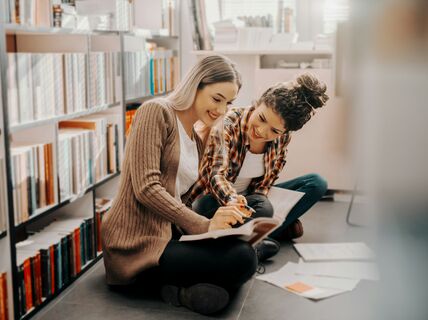 Two people sitting on the floor in a library, looking at a book. Papers are scattered around them.