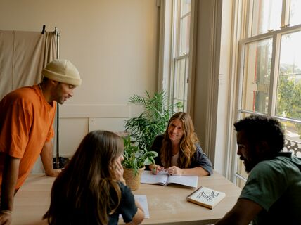 Four people collaborating at a table with notebooks and a plant, next to a window with natural light.