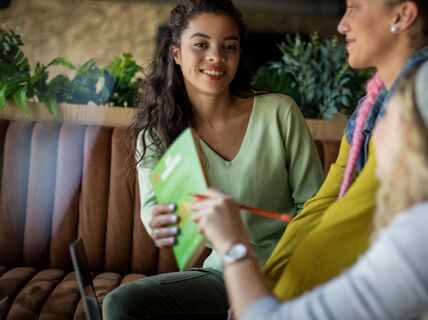 Three people sitting on a cushioned bench in a cosy cafe, one holding a green menu, surrounded by green plants.
