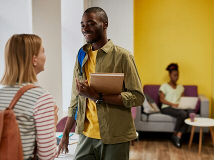 Två personer pratar tillsammans. En håller en anteckningsbok. En person sitter i soffan i bakgrunden. 