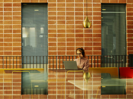 Henkilö työskentelee kannettavalla pöydän ääressä, tiiliseinä ja raidalliset ikkunat heijastuvat pöytään.