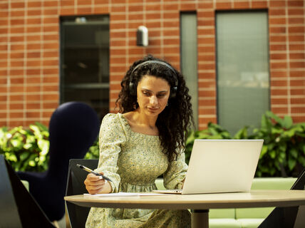 Henkilö, jolla on kiharat hiukset ja kuulokkeet, työskentelee kannettavalla tietokoneella ulkona ja pitelee kynää. Taustalla on tiiliseinä ja kasveja.