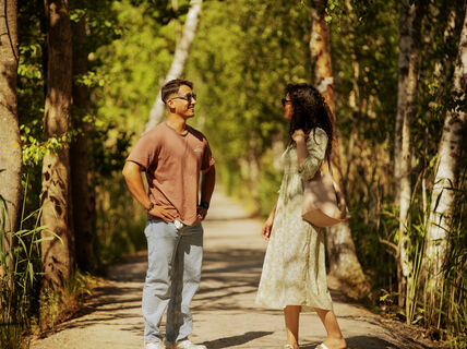 Two people talking on a sunlit forest path, one in a brown shirt and jeans, the other in a light dress and sandals.