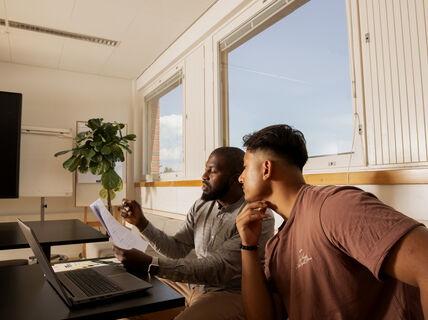 Two people in a room with large windows, one holding papers and the other looking at them. A laptop is on the table.