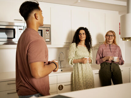 Kolme ihmistä seisoo työpaikan keittiössä pitäen mukeja, taustalla on pesuallas ja mikroaaltouuni.