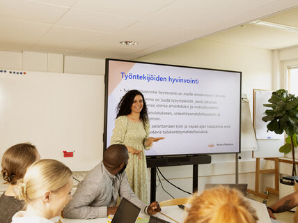 Henkilö esittelee ryhmälle kokoushuoneessa. Näytöllä lukee Työntekijöiden hyvinvointi