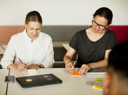 Two people seated at a table writing on sticky notes with a laptop and a phone on the table.