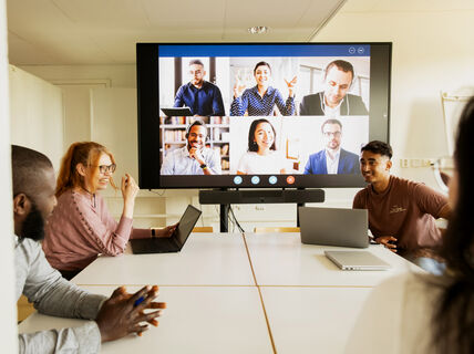 Människor i ett mötesrum deltar i en videokonferens. Stor skärm visar fler personer på samtalet.
