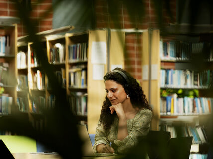 A person with curly hair is sitting in a library, wearing headphones and using a laptop. Bookshelves are in the background.