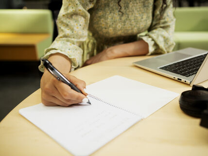 En person skriver i en anteckningsbok på ett träbord med en laptop och hörlurar i närheten.