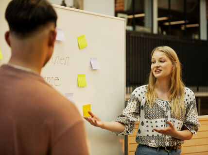 Kaksi henkilöä keskustelemassa ulkona, valkotaululla on muistilappuja. Nainen elehtii käsillään.