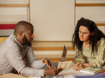 Kaksi ihmistä istuu pöydän ääressä, jossa on kannettava tietokone ja kirjoja. He keskustelavat paperista.