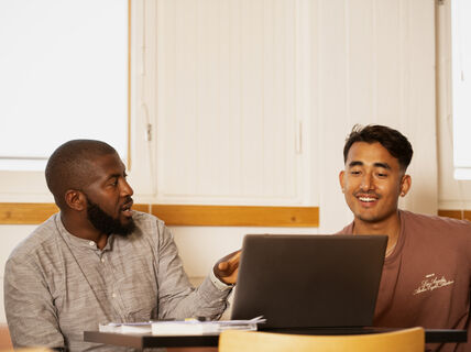 Two individuals are sitting at a table with a laptop, discussing something in a brightly lit room.