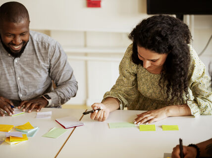 Två personer sitter vid ett bord med klisterlappar, pennor och en anteckningsbok, engagerade i en samarbetsuppgift.