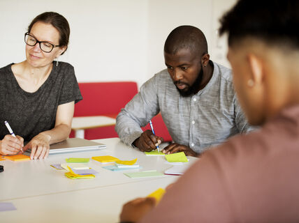 Kolme ihmistä tekee yhteistyötä pöydän ääressä, jossa on muistilappuja ja kannettavia tietokoneita.