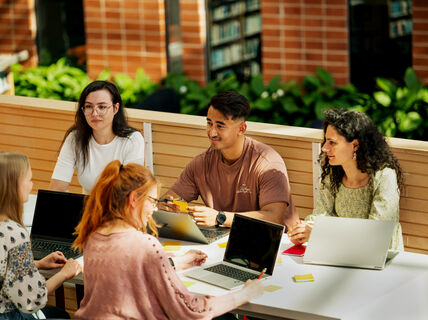 En grupp människor som sitter runt ett bord med bärbara datorer och anteckningsböcker i en ljus inomhusmiljö med växter och bokhyllor.