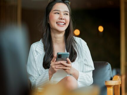 Pitkähiuksinen henkilö istuu sisällä käyttämässä älypuhelinta. Hänellä on yllään vaalea raidallinen paita.