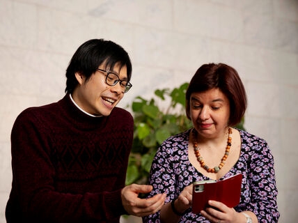 Two people are looking at a red phone. One person is wearing a maroon sweater, and the other a floral top and beaded necklace.