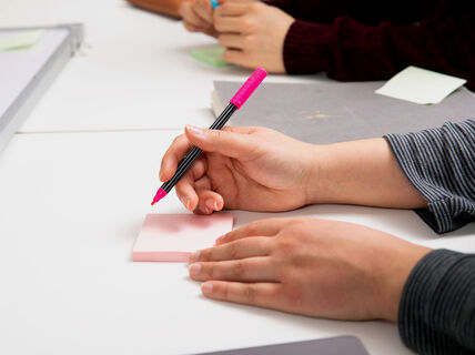 Lähikuva käsistä kirjoittamassa vaaleanpunaisella kynällä muistilapulle valkoisen pöydän ääressä, taustalla muita lappuja ja ihmisiä.