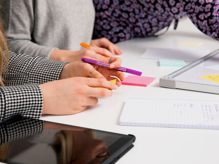 Ihmiset työskentelevät pöydän ääressä muistikirjojen, muistilappujen ja tabletin kanssa. Yksi pitelee violettia kynää.
