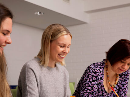 Kolme henkilöä istuu pöydän ääressä, kaksi katsoo tarkasti papereita. Huoneessa on valkoiset seinät ja moderni sisustus.