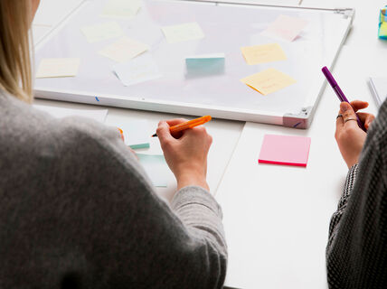 Kaksi ihmistä kirjoittaa muistilappuihin työpöydällä, taustalla valkotaulu ja lisää muistilappuja.
