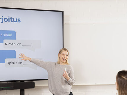 Henkilö osoittaa suurta näyttöä, jossa on teksti 'Harjoitus', ja pitelee tussia. Toinen henkilö etualalla.