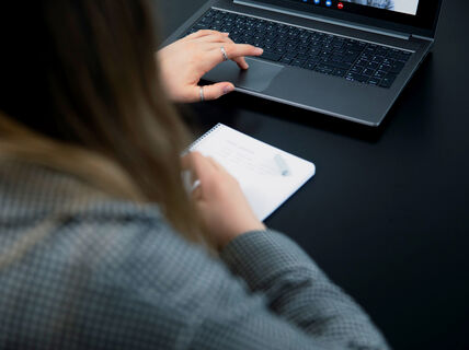 Henkilö kirjoittaa muistikirjaan kynällä ja käyttää kannettavaa tietokonetta mustan pöydän ääressä videopuhelussa.