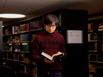Person in a red sweater reading a book in a library aisle. Shelves are filled with various coloured books.