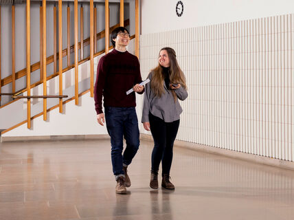 Two people walking indoors. The man holds a notebook and wears a red jumper. The woman holds a phone and wears a grey top.
