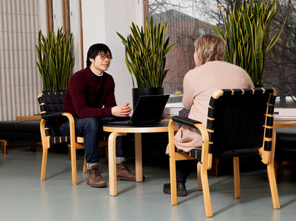 Kaksi ihmistä istuu pienen pöydän ääressä modernissa huoneessa, jossa on isot ikkunat, kasveja ja puuhuonekaluja. Toisella on kannettava tietokone auki.