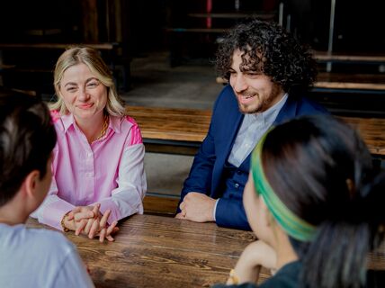 Four people are sitting at a wooden table, having a conversation. Two have their backs to the camera.