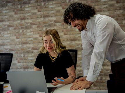 Two people in an office, one seated at a laptop showing something to the other who is standing and leaning in.
