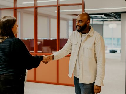 Kaksi henkilöä kättelee toimistossa, taustalla on punaiset väliseinät ja työpöytiä.