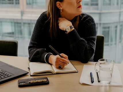 Henkilö kirjoittaa muistivihkoon pöydällä, jossa on kannettava tietokone, puhelin, kynä, paperi ja vesilasi.