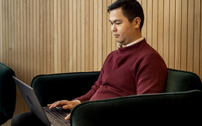 A person in a red sweater sits on a chair working on a laptop. Wooden panel wall in the background.