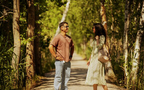Två personer pratar på en solbelyst skogsstig, en i brun skjorta och jeans, den andra i ljus klänning och sandaler.