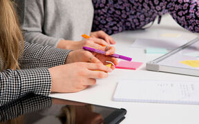 People working at a table with notebooks, sticky notes, and a tablet. One holds a purple pen.