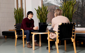 Kaksi ihmistä istuu pienen pöydän ääressä modernissa huoneessa, jossa on isot ikkunat, kasveja ja puuhuonekaluja. Toisella on kannettava tietokone auki.