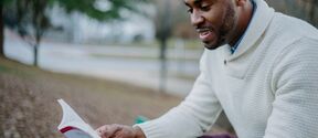 A person wearing a white jumper sits on a park bench, reading a book with a red and white cover.
