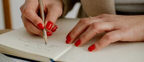 Henkilö, jolla on punaista kynsilakkaa, kirjoittaa muistikirjaan, yllään beige bleiseri.