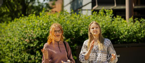Two people are standing outside near some greenery, one in a pink top and the other in a patterned white top.