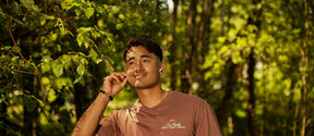 A person wearing a brown T-shirt stands in a forested area, with their hand touching a leafy branch.