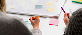 Kaksi ihmistä kirjoittaa muistilappuihin työpöydällä, taustalla valkotaulu ja lisää muistilappuja.