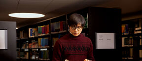 Person in a red sweater reading a book in a library aisle. Shelves are filled with various coloured books.
