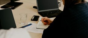 Henkilö kirjoittaa muistikirjaan pöydällä, jossa on kannettava tietokone, vesilasi, tabletti ja kynä.