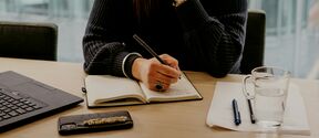 A person writes in a notebook at a desk with a laptop, phone, pen, paper, and glass of water.