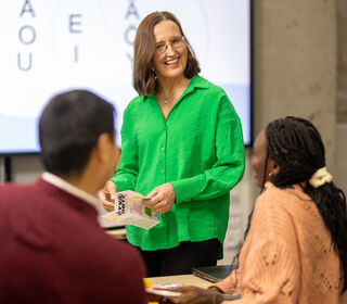 En lärare i en grön skjorta instruerar två elever i ett klassrum med en whiteboard som visar vokaler.