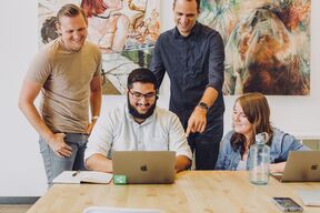 Four people in a meeting room with laptops on a wooden table and colourful paintings on the wall.