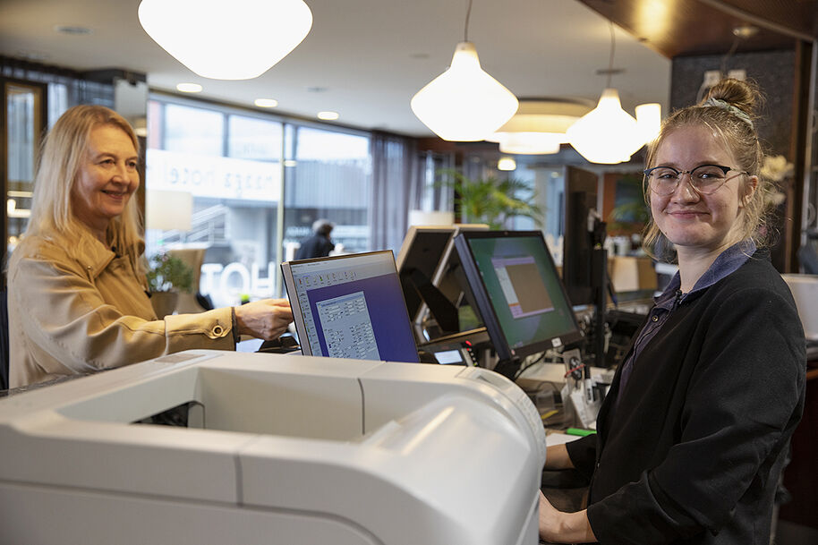 Kuva on hotellin vastaanotosta. Kuvassa näkyvät asiakas ja vastaanottovirkailija. Virkailija katsoo kmameraan ja hymyilee. 